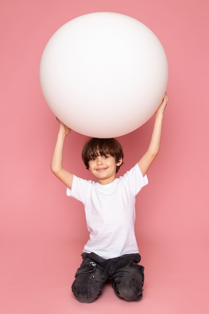 Een jongen van het vooraanzicht glimlachende kind in het witte t-shirt spelen met witte bal op de roze ruimte