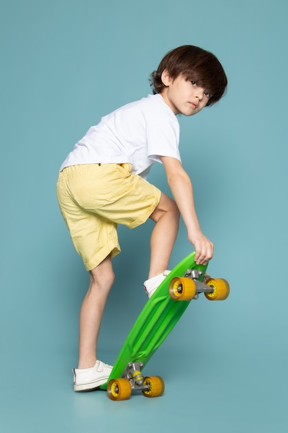 Een jongen van het vooraanzichtkind in witte t-shirt en gele jeans die groen skateboard berijden op de blauwe ruimte