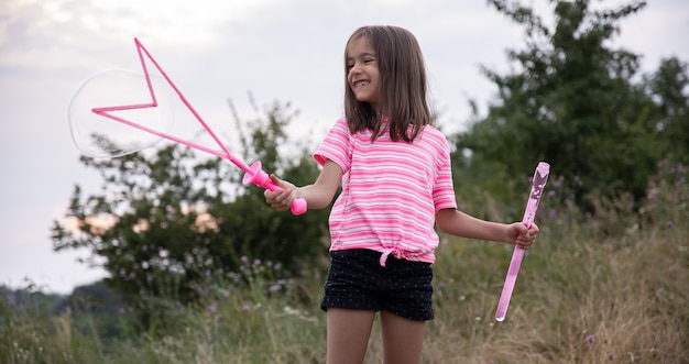Een klein grappig meisje blaast zeepbellen in de zomer in een veld, buiten zomeractiviteiten.