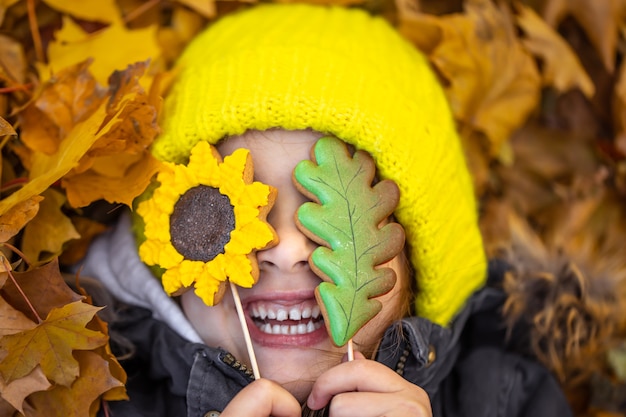 Gratis foto een klein grappig meisje met een gele hoed ligt in het herfstgebladerte en houdt peperkoek in haar handen.