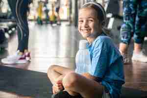 Gratis foto een klein meisje met een fles water zit op een mat in de sportschool