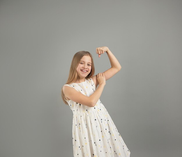 Een klein meisje toont haar kracht. een mooi jong meisje dat de kracht van haar armen laat zien, poseert in een lichte lichte jurk in de studio op een grijze achtergrond.