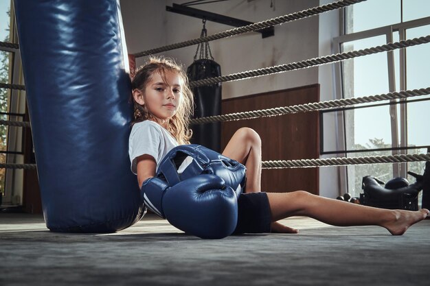 Een klein vermoeid meisje met een blauwe helm rust op de ring naast de bokszak.