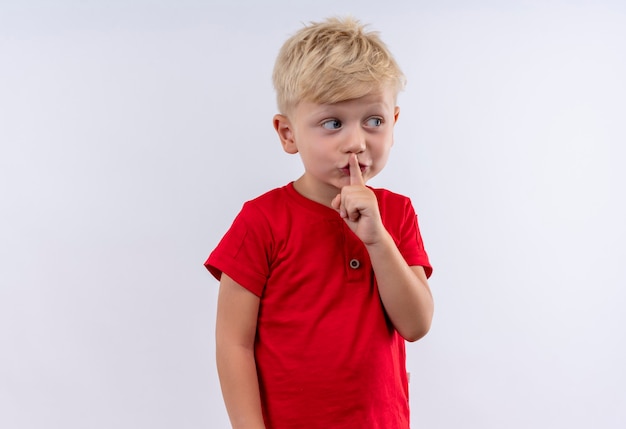 Een kleine schattige blonde jongen in rood t-shirt toont shh gebaar met wijsvinger op zijn mond terwijl hij kant op een witte muur kijkt