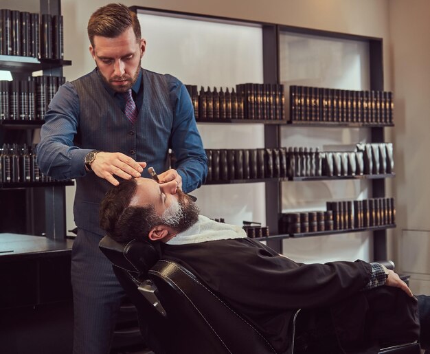 Een knappe hipster bebaarde man zittend in een fauteuil in een kapperszaak terwijl kapper zijn baard scheert met een gevaarlijk scheermes.