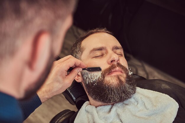 Een knappe hipster bebaarde man zittend in een fauteuil in een kapperszaak terwijl kapper zijn baard scheert met een gevaarlijk scheermes.