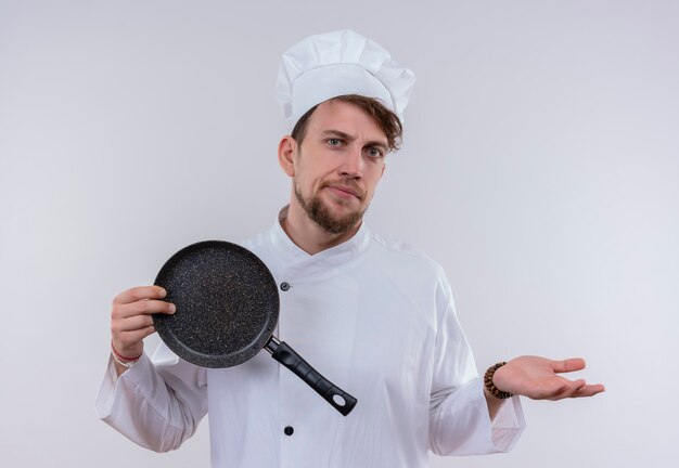 Een knappe jonge bebaarde chef-kok man met een wit fornuis uniform en een hoed die koekenpan toont terwijl hij met verbaasde uitdrukking op een witte muur kijkt