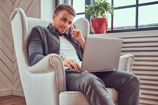 Een knappe jonge kerel in vrijetijdskleding die op zijn laptop werkt terwijl hij op een stoel zit.