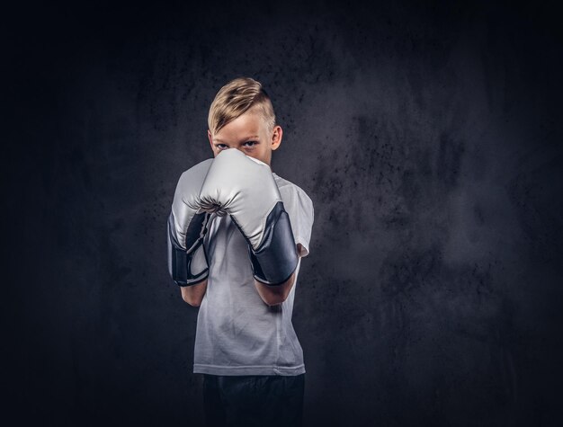 Een knappe kleine jongensbokser met blond haar, gekleed in een wit t-shirt in handschoenen, klaar om te vechten. Geïsoleerd op de donkere gestructureerde achtergrond.