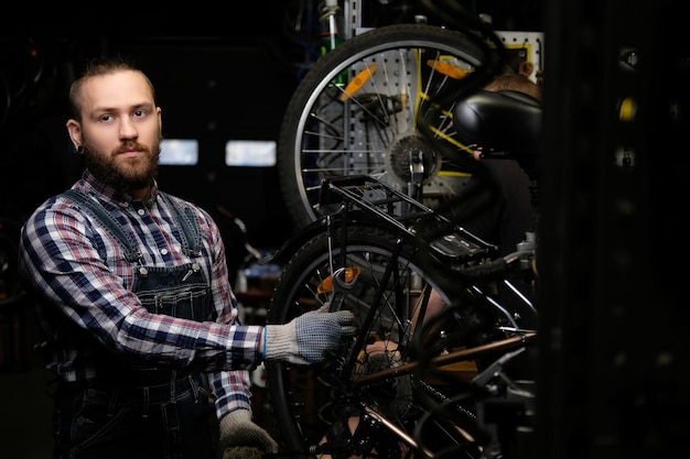Een knappe stijlvolle man met een flanellen hemd en jeans overall, werkend met een fietswiel in een reparatiewerkplaats. Een arbeider die een moersleutel gebruikt, monteert het wiel op een fiets in een werkplaats.