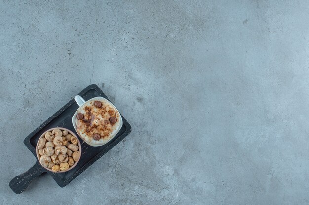 Een kom cornflakes en een kopje cappuccino op een bord, op de blauwe achtergrond.