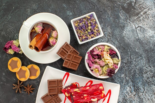 Een kopje kruidenthee met droge bloemen en koekjes en een bord chocolaatjes op grijze grond