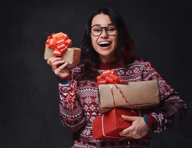 Een lachende brunette vrouw gekleed in een rode trui en bril houdt kerstcadeaus over grijze achtergrond.