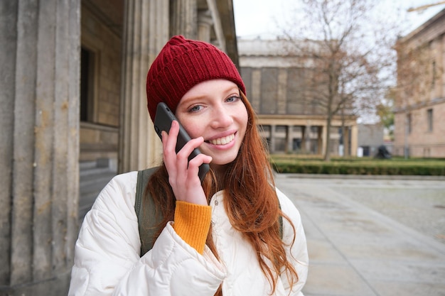 Gratis foto een lachende jonge roodharige vrouw luistert naar een spraakbericht, maakt een telefoontje, loopt op straat en praat met