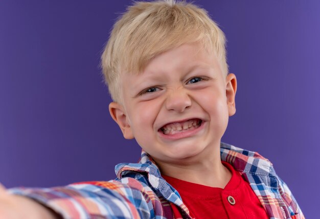 Een lachende schattige kleine jongen met blond haar en blauwe ogen, gekleed in geruit overhemd kijken op een paarse muur