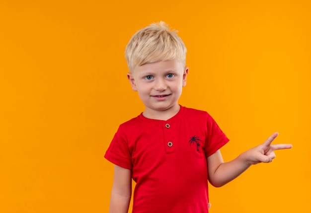 Een lachende schattige kleine jongen met blond haar, gekleed in een rood t-shirt met twee vingers gebaar op een gele muur