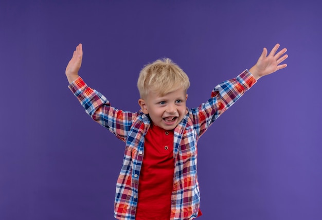 Een lachende schattige kleine jongen met blond haar in een geruit overhemd met zijn handen in de lucht terwijl hij op een paarse muur kijkt