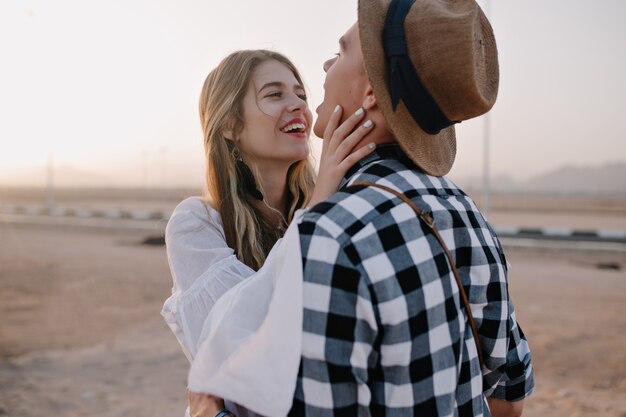 Een lachende vrouw in een wit overhemd streelt haar vriendje in het gezicht en kijkt met liefde naar zijn ogen. Jonge man met een geruit overhemd tijd doorbrengen met vriendin op romantische date buiten in de ochtend