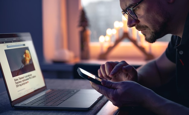 Een man met een smartphone zit 's avonds laat voor een laptop