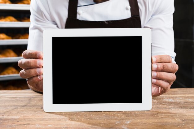 Een mannelijke bakker in uniform die kleine lege digitale tablet op houten lijst houdt