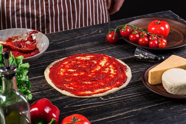 Een mannenhand verspreiding van tomatenpuree op een pizzabodem met lepel op een oude houten achtergrond. Koken concept. Detailopname