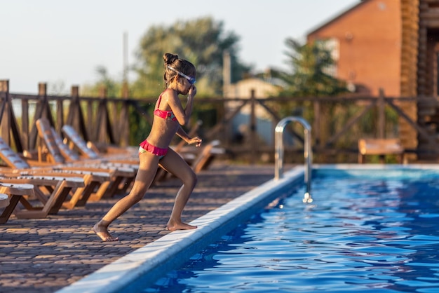 Een meisje in felroze zwembroek en zwarte zwembril, grappig springend in diepblauw zwembad met schoon transparant koel water tegen de achtergrond van de warme zomerzonsondergang