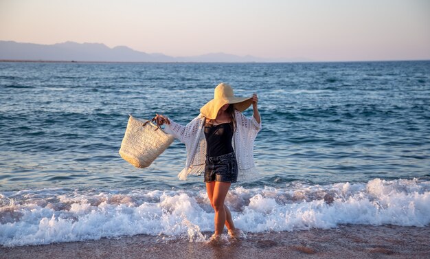 Een meisje met een grote hoed en een rieten tas loopt langs de zeekust. Zomer vakantie concept.