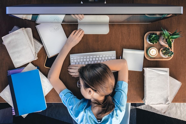 Een moe meisje ligt op een tafel voor een computer met notitieblokken en boeken