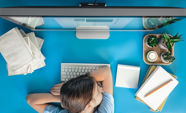 Een moe meisje ligt op een tafel voor een computer met notitieblokken en boeken