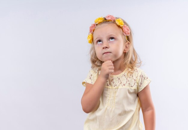 Een mooi klein meisje draagt een geel shirt in een bloemenhoofdband en denkt met de hand op de kin en kijkt omhoog op een witte muur