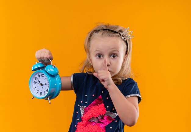 Een mooi klein meisje met een marineblauw shirt in een hoofdband met een kroon dat haar gebaar toont terwijl ze een blauwe wekker op een oranje muur houdt