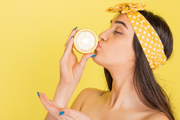 Een mooi meisje dat plastic sinaasappelschijfje kust en op gele achtergrond staat