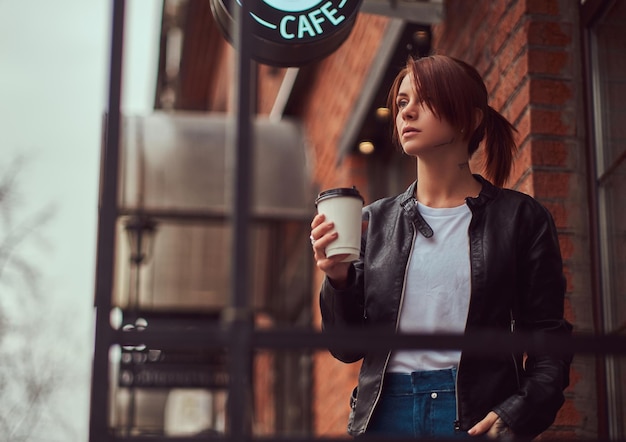 Een mooi meisje draagt een leren jas met een rugzak die een beker vasthoudt met afhaalkoffie buiten in de buurt van het café.