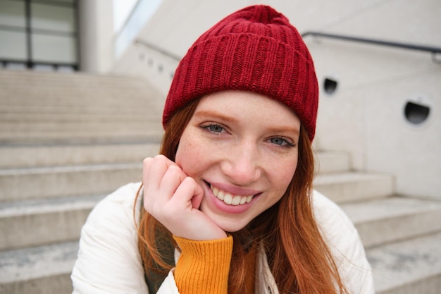 Gratis foto een mooi roodharig studentenmeisje met een rode hoed glimlacht oprecht en ziet er gelukkig en ontspannen uit op de trap