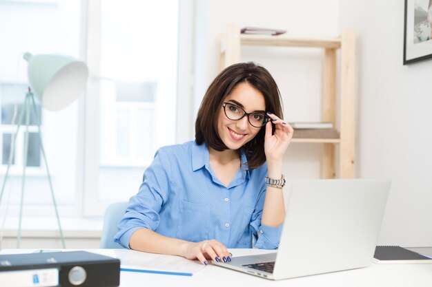 Een mooie brunette meisje in blauw shirt zit aan de tafel op kantoor. Ze werkt met laptop. Ze lacht naar de camera.