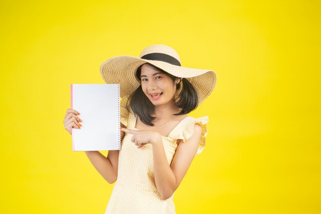 Een mooie, gelukkige vrouw die een grote hoed draagt en een wit boek op een geel houdt.