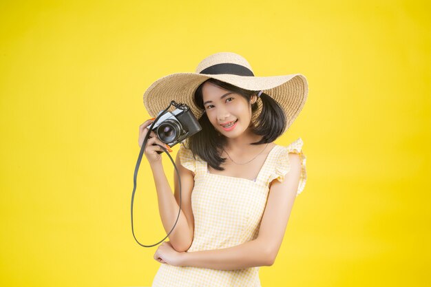 Een mooie, gelukkige vrouw die een grote hoed en een camera op een geel draagt.