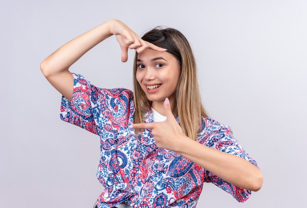 Een mooie jonge vrouw die een paisley bedrukt overhemd draagt en een frame met handen en vingers maakt met een blij gezicht terwijl ze op een witte muur kijkt