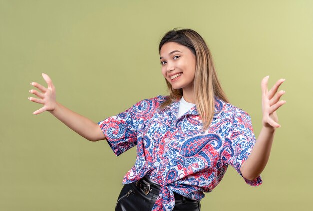 Een mooie jonge vrouw die een paisley-bedrukt overhemd draagt en haar handen opent om te knuffelen terwijl ze op een groene muur kijkt