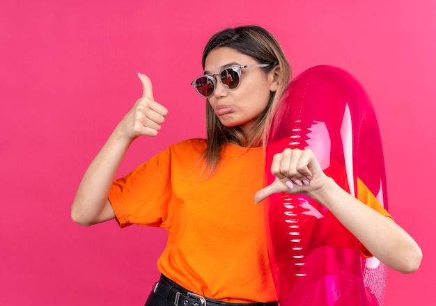 Een mooie jonge vrouw in een oranje t-shirt met een zonnebril die duimen op en neer laat zien terwijl ze een roze opblaasbare ring vasthoudt aan een roze muur