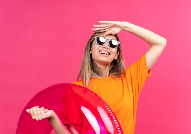 Een mooie jonge vrouw in een oranje t-shirt met een zonnebril die glimlacht en ver weg kijkt terwijl ze een roze opblaasbare ring op een roze muur vasthoudt