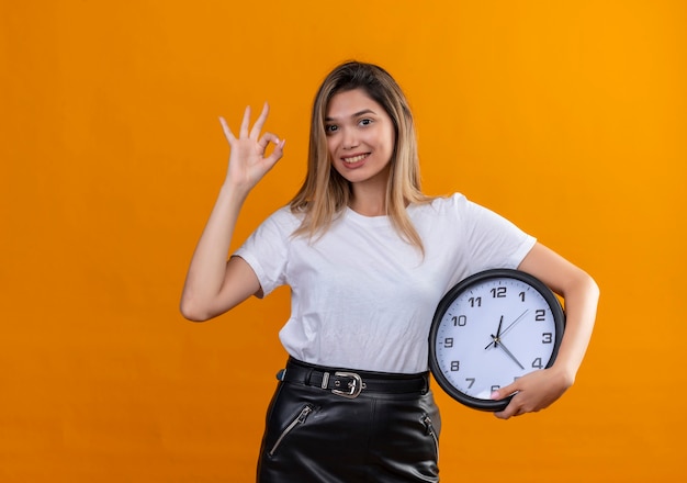 Een mooie jonge vrouw in een wit t-shirt die ok teken toont terwijl zij een muurklok op een oranje muur houdt