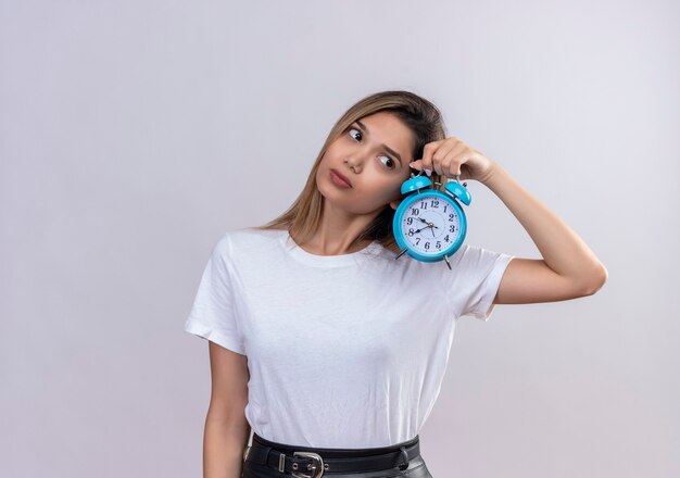 Een mooie jonge vrouw in wit t-shirt luistert naar het tikkende geluid van de klok terwijl ze een blauwe wekker op een witte muur houdt