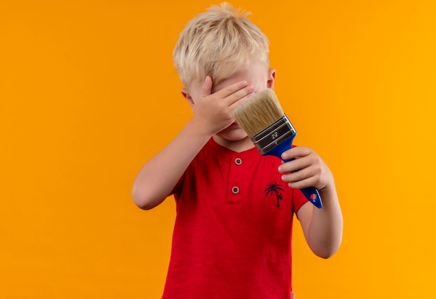 Een mooie kleine jongen met blond haar, gekleed in een rood t-shirt met blauwe verfborstel met hand bedekkende ogen op een gele muur