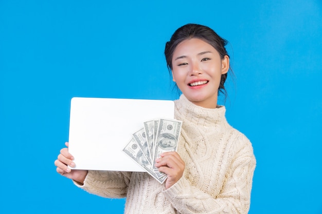 Een mooie vrouw die een nieuw wit tapijt met lange mouwen draagt dat een wit teken en een dollarrekening op een blauw houdt. Handel.