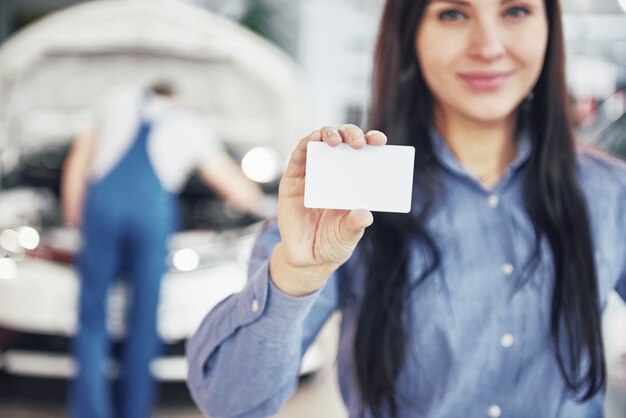 Een mooie vrouw houdt een visitekaartje van het autoservicecentrum. De monteur inspecteert de auto onder de motorkap op de achtergrond