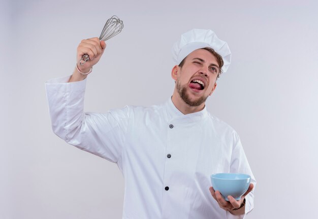 Een ontevreden jonge bebaarde chef-kok man met wit fornuis uniform en hoed verhogen mixer lepel met blauwe kom in de andere hand op een witte muur