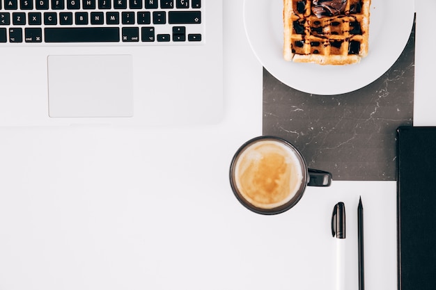 Een open laptop; wafel; koffiekop; potlood; pen en open laptop op witte bureau