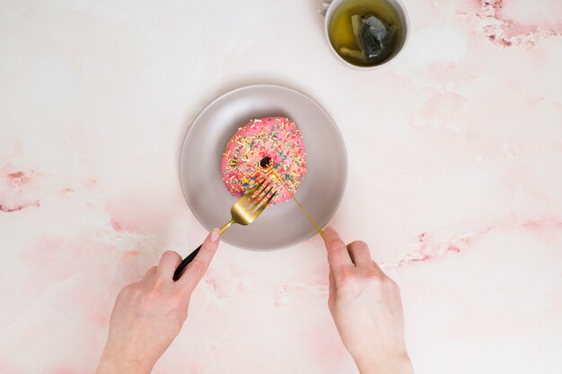 Een opgeheven mening van een persoon die de donuts met vork en botermes snijdt op geweven achtergrond