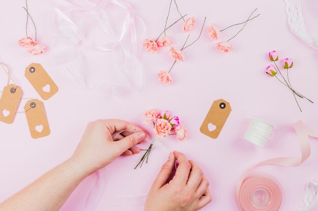 Een opgeheven mening van menselijke hand die de kunstbloemen met lint op roze achtergrond bindt